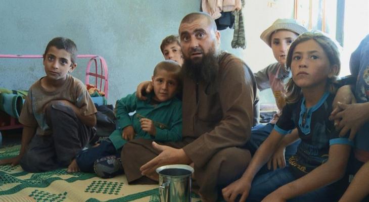 A group of people sitting on the floor looking at the camera. One man with a beard and a bunch of children.