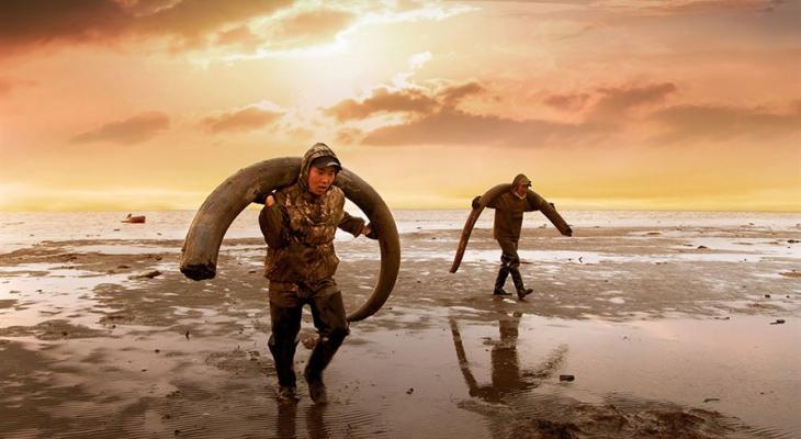 Two men walking through mud carrying mammoth tusks
