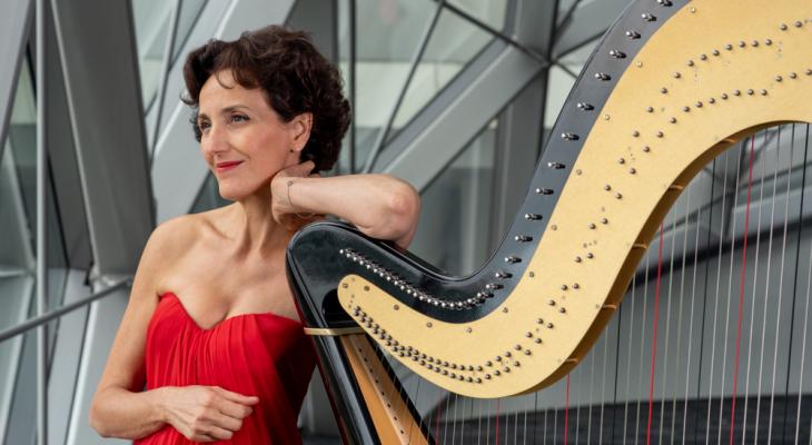 Isabelle Moretti, a woman with short, curly, dark brown hair and brown eyes, stands in front of a glass wall, with her elbow resting on her harp. She is wearing a red dress and red lipstick, and smiling.