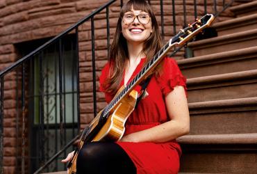 Jocelyn Gould is sitting on the steps, smiling, wearing a red dress, and holding a jazz guitar in her hand.
