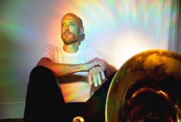 Tom Richards sits on the ground leaning against a white wall, gazing into the distance. He is wearing a white T-shirt and black pants, with colorful halos of light around him.