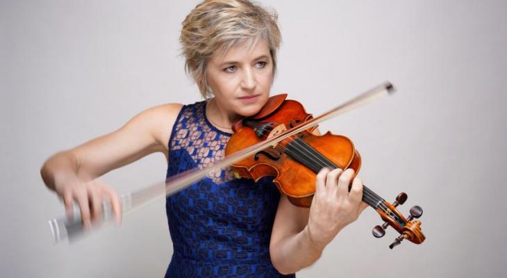 Marianne Piketty standing in front of a grey background, dressed in a dark blue dress, and playing a violin.