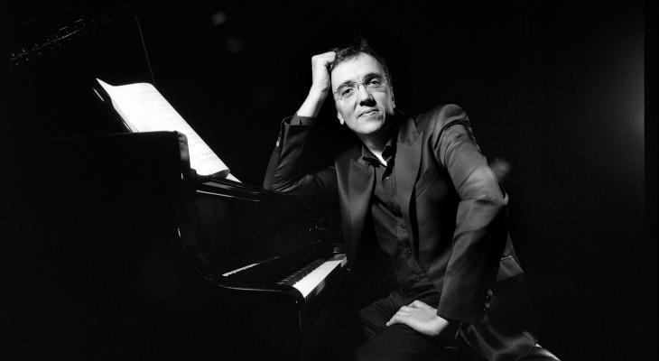 Éric Le Sage is sitting at the piano, leaning on the instrument and smiling at the camera.