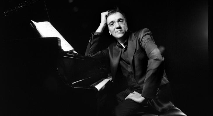 Éric Le Sage is sitting at the piano, leaning on the instrument, and looking at the camera, smiling.