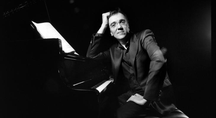 Éric Le Sage is sitting at the piano, leaning on the instrument and looking at the camera, smiling.