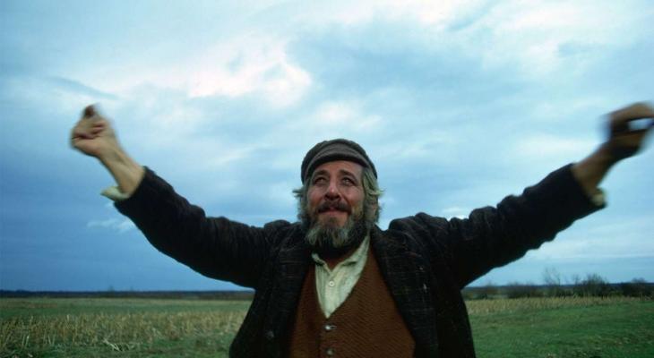 Tevye stands in grassy field with his arms stretched to the sky.