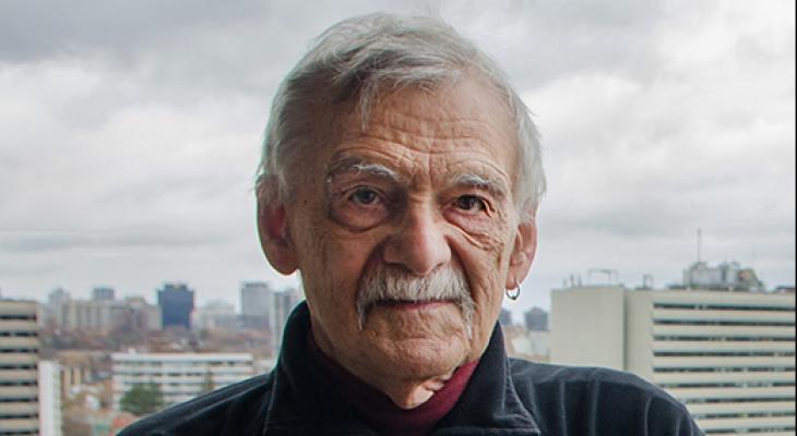 An elderly man poses for a portrait, with the backdrop of a city behind him
