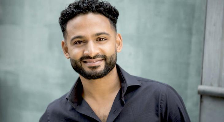 Conductor Jordan de Souza is smiling at the camera. He has brown skin, dark curly hair and a beard and is wearing a black long-sleeve shirt.