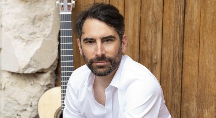 Jérémy Jouve wearing a white dress shirt, sitting on a rustic threshold with his guitar behind him.