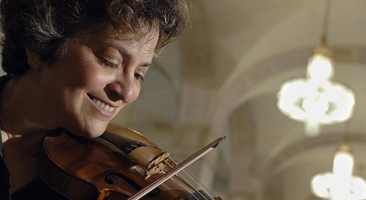 Close-up portrait of Jeanne Lamon, smiling, playing her violin in a large room with arched walls and bright chandeliers.