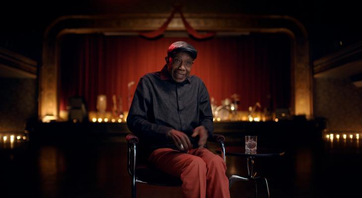 A man seated in a darkened theatre addresses the camera.