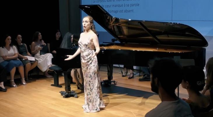 A singer is singing in the foreground with a grand piano behind her.