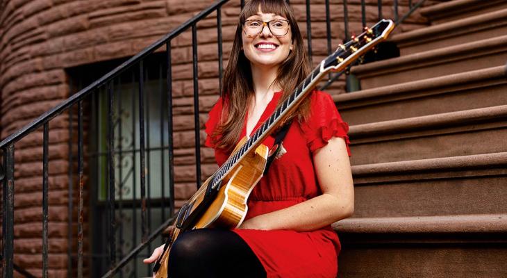 Jocelyn Gould is sitting on the steps, smiling, wearing a red dress, and holding a jazz guitar in her hand.