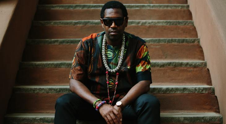 Yosvany Terry is seated on steps looking straight at the camera. He is wearing sunglasses, a multicoloured shirt and dark pants, and beaded jewellery around his neck and wrist.