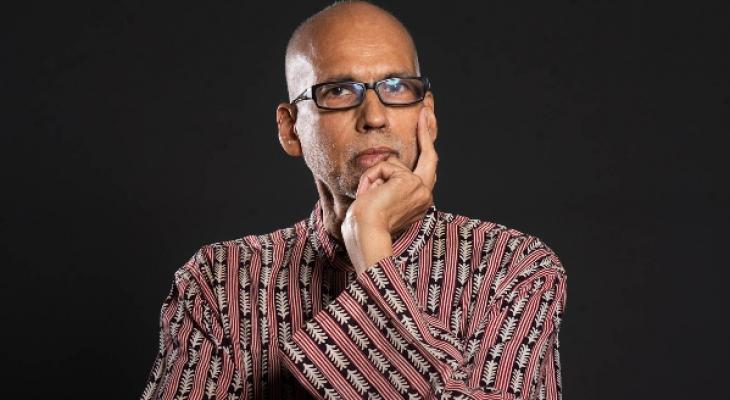 André de Quadros stands in front of a black background, wearing black-framed glasses, a floral shirt, with one hand resting on his face.