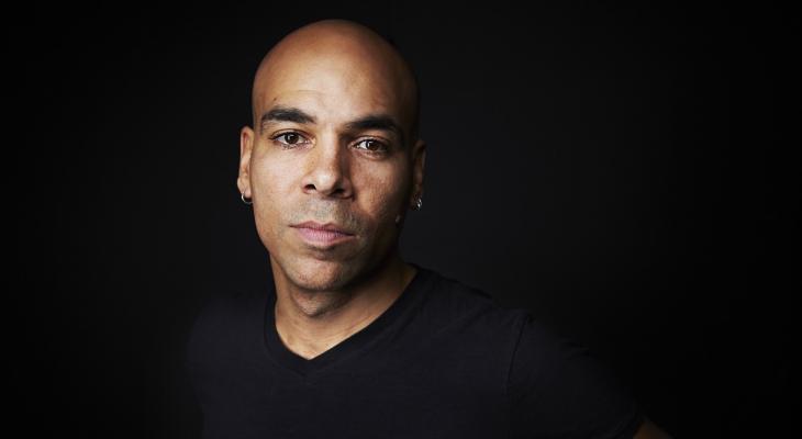 Actor/musician Beau Dixon is pictured in front of a dark background. He is a Black man with a shaved head and is facing the camera wearing a black t-shirt.