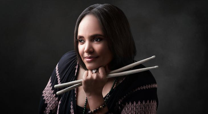 Terri Lyne Carrington is pictured against a grey background, looking away from the camera, her head resting on her fist holding a pair of drumsticks. She is a Black woman with straight, shoulder-length hair and is wearing a black and pink patterned shirt.