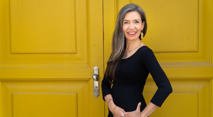 Patricia Caicedo standing in front of a yellow door, smiling, wearing a black dress, with her hair down.