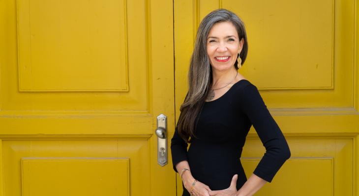 Patricia Caicedo standing in front of a yellow door, smiling, wearing a black dress, with her hair down.
