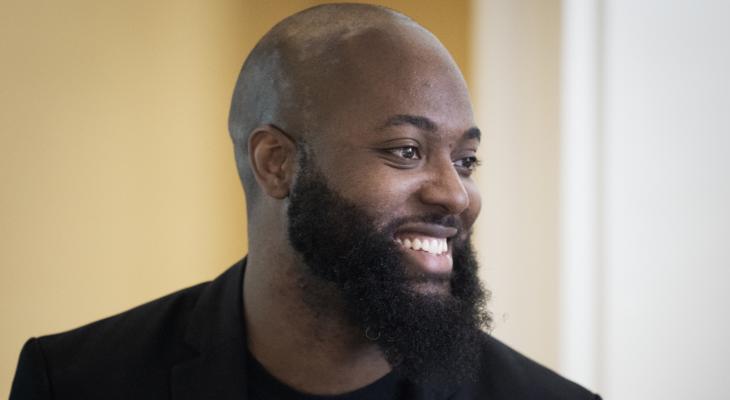 Nathan Holder is smiling and looking to the right. He has a beard and is wearing a black suit jacket over a black T-shirt with white text printed on it.