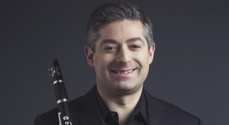 Peter Stoll is wearing a black suit, with a black background behind him. He is smiling and looking ahead, holding a clarinet in his hand.
