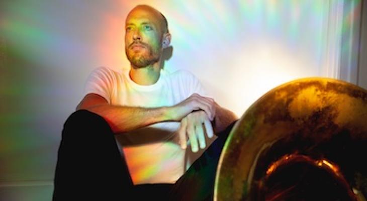 Tom Richards sits on the ground leaning against a white wall, gazing into the distance. He is wearing a white T-shirt and black pants, with colorful halos of light around him.