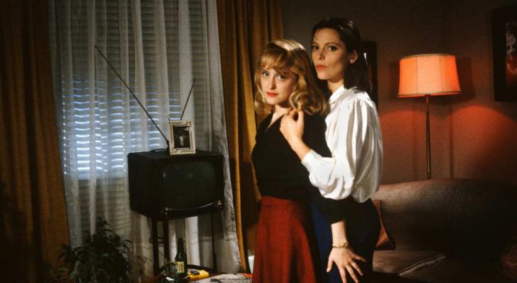 Two lesbians embrace in a 1950s-style Canadian living room.
