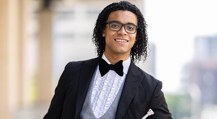William Leathers outdoors, dressed in a black suit, wearing a bowtie, glasses, holding a trumpet, smiling at the camera.