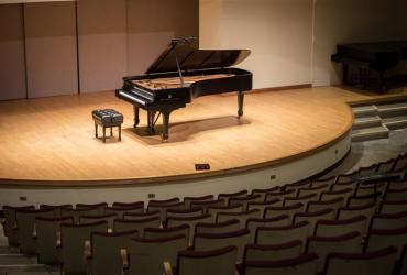 A grand piano sits in the spotlight on the Walter Hall stage surrounded by empty seats.