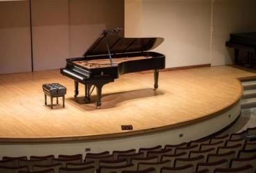 A grand piano sits in the spotlight on the Walter Hall stage surrounded by empty seats.