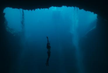 Floating deep in an underwater cavern, a free-diver extends her arms towards the surface.