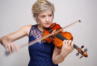 Marianne Piketty standing in front of a grey background, dressed in a dark blue dress, and playing a violin.