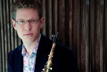 Wallace Halladay leans against a wooden background wall. He is wearing a black suit jacket and a floral shirt, holding a saxophone in his hand.