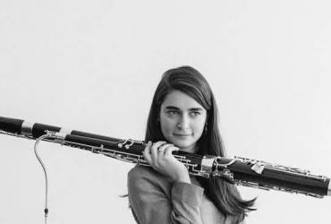 Black and white image of Sophie Dervaux, a woman with long, dark hair, holding a bassoon over her shoulder.