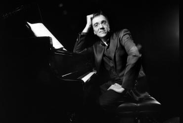 Éric Le Sage is sitting at the piano, leaning on the instrument, and looking at the camera, smiling.