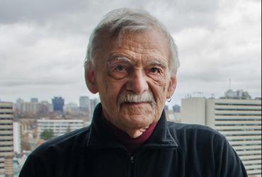 An elderly man poses for a portrait, with the backdrop of a city behind him 