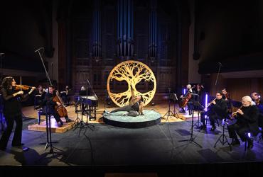 An actor poses in front of a wooden carving of a maple tree, centre stage under a bright light, with Tafelmusik musicians performing around them.