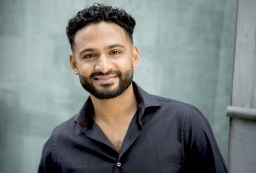 Conductor Jordan de Souza is smiling at the camera. He has brown skin, dark curly hair and a beard and is wearing a black long-sleeve shirt.