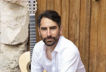 Jérémy Jouve wearing a white dress shirt, sitting on a rustic threshold with his guitar behind him. 