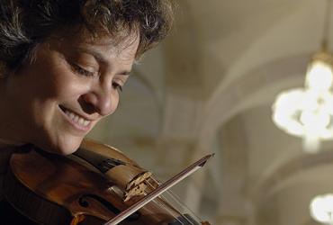 Close-up portrait of Jeanne Lamon, smiling, playing her violin in a large room with arched walls and bright chandeliers.