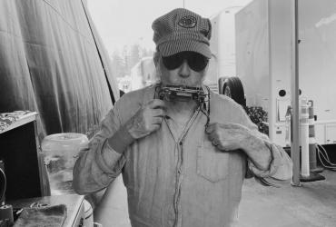 Neil Young with a striped cap and sunglasses playing harmonica