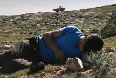 A man with a video camera lies in the sun on a rocky hillside.