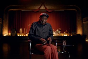 A man seated in a darkened theatre addresses the camera.