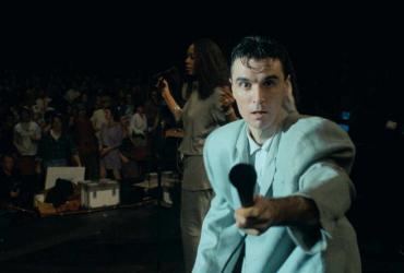 David Byrne reaches out a microphone towards the camera.