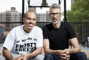 DJs Stretch and Bobbito smile for the camera outside a basketball court. 