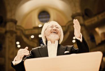 Close-up of Masaaki Suzuki conducting from the podium. He is an older Asian male, smiling and wearing a black tuxedo and white white with a high collar.