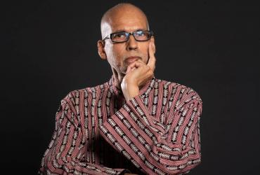André de Quadros stands in front of a black background, wearing black-framed glasses, a floral shirt, with one hand resting on his face.