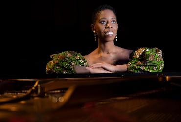 Dr. Ege is seated, leaning on a grand piano, smiling and looking into the distance. She is a Black woman wearing a green, yellow and black patterned top and dangling earrings.