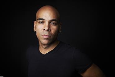 Actor/musician Beau Dixon is pictured in front of a dark background. He is a Black man with a shaved head and is facing the camera wearing a black t-shirt.
