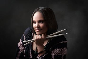 Terri Lyne Carrington is pictured against a grey background, looking away from the camera, her head resting on her fist holding a pair of drumsticks. She is a Black woman with straight, shoulder-length hair and is wearing a black and pink patterned shirt.